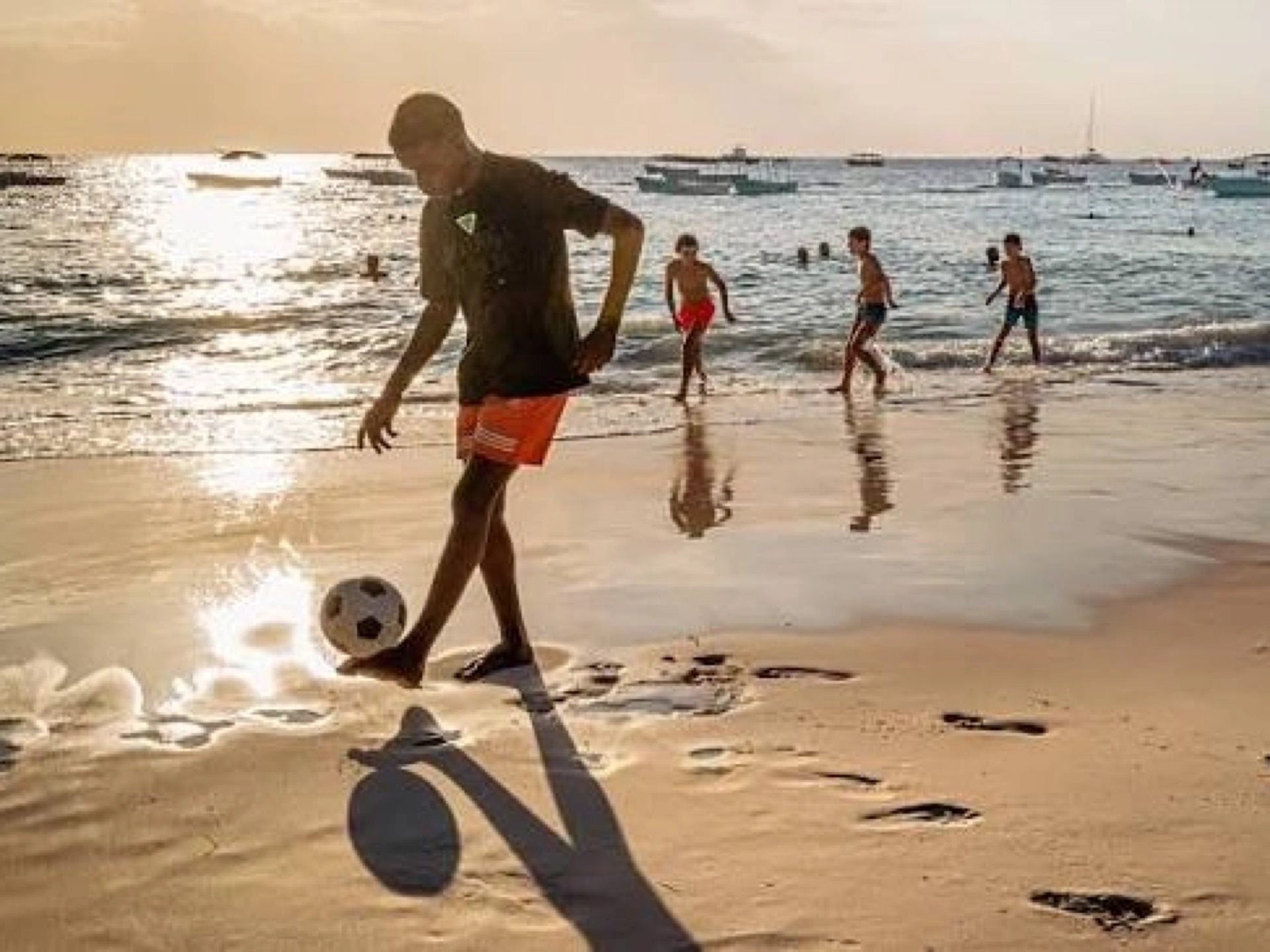 A man playing football with children at the water’s edge on a Zanzibar beach at sunset, with dhows anchored offshore